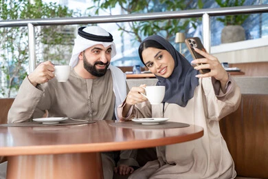 التحدث والتواصل مع العائلة والأصدقاء، احتساء المشروب المفضل، إجراء مكالمات الفيديو عبر الانترنت، استخدام اجهزة تقنية حديثة، رجل عربي خليجي اماراتي يرتدي الكندورة و الغتره يجلس فى احد المقاهي برفقة زوجته ، ايماءات السعادة و السرور