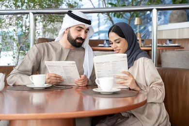 Choosing meals and drinks from the restaurant menu, having a good time at the café, an Emirati Arab couple in traditional attire sitting in one of the cafés exchanging conversation, a happy family atmosphere.