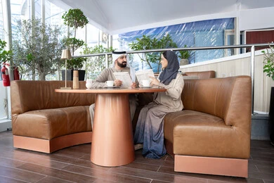 اختيار الأطباق والمشروبات من قائمة الطعام، قضاء وقت ممتع في المقهى، زوجان عربيان خليجيان إماراتيان يرتديان الزي التقليدي يجلسان في أحد المقاهي يتبادلان الحديث، اجواء عائلية سعيده