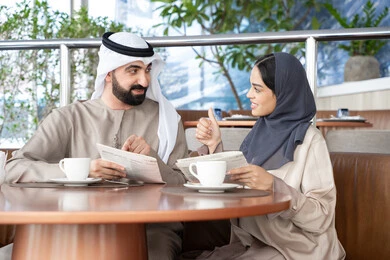 إيماءات يدين تدل على الإعجاب والرضى، اجواء عائلية سعيده، قضاء وقت ممتع في الكافية، اختيار الأطباق والمشروبات من قائمة الطعام، زوجان عربيان خليجيان إماراتيان يرتديان الزي التقليدي يجلسان في أحد المقاهي يتبادلان الحديث