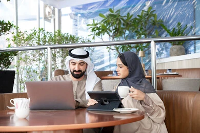 الاستمتاع بتناول المشروبات المفضله، إنجاز الأعمال المطلوبة، دمج التقنية والتكنولوجيا في الروتين اليومي، مفهوم العمل عن بعد، ايماءات التركيز والسرور، قضاء وقت ممتع في الخارج، زوجان عربيان خليجيان اماراتيان يرتديان الزي التقليدي يجلسان في احد المقاهي يعملان على الحاسب المحمول وجهاز التابلت