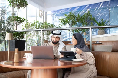 إنجاز الأعمال المطلوبة، الاستمتاع بتناول المشروبات المفضله، دمج التقنية والتكنولوجيا في الروتين اليومي، مفهوم العمل عن بعد، ايماءات التركيز والسرور، قضاء وقت ممتع في الخارج، زوجان عربيان خليجيان اماراتيان يرتديان الزي التقليدي يجلسان في احد المقاهي يعملان على الحاسب المحمول وجهاز التابلت