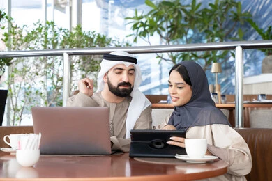 The concept of remote work, gestures of focus and joy, spending enjoyable time outdoors, accomplishing daily tasks, integrating technology and tech into the daily routine, an Emirati Arab couple in traditional attire sitting in a café working on a laptop and a tablet.