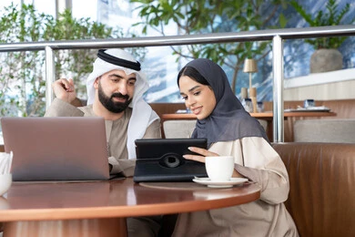 Spending enjoyable time outdoors, gestures of focus and joy, remote work, accomplishing daily tasks, integrating technology and tech into the daily routine, an Emirati Gulf Arab couple dressed in traditional attire sitting in a café working on a laptop and a tablet.
