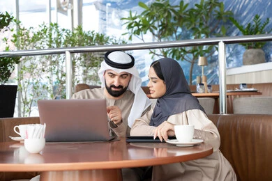 Managing and organizing business affairs through modern technological devices, dialogue and discussion among colleagues, using a laptop to accomplish projects and tasks in the café, an Emirati Gulf Arab man wearing a kandura and ghutrah sitting with his female colleague in one of the cafés, expressions of happiness and joy.