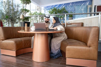 الاستمتاع بالوقت مع الأصدقاء، الحوار والنقاش بين زملاء العمل، استخدم جهاز الحاسوب المحمول لانجاز المشاريع والمهام في الكافيه, رجل عربي خليجي اماراتي يرتدي الكندورة و الغتره يجلس مع زميلته فى احد المقاهي ، تعابير السعادة و السرور