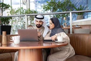 الحوار والنقاش بين زملاء العمل، استخدم جهاز الحاسوب المحمول لانجاز المشاريع والمهام في الكافيه، قضاء وقت ممتع مع الأصدقاء, رجل عربي خليجي اماراتي يرتدي الكندورة و الغتره يجلس مع زميلته فى احد المقاهي ، تعابير السعادة و السرور