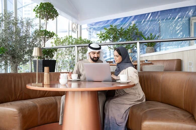 The dialogue and discussion among colleagues at work, using a laptop to complete projects and tasks in the café, spending enjoyable time with friends, an Arab Gulf Emirati man wearing a kandura and ghutrah sitting with his female colleague in one of the cafés, expressions of happiness and joy.
