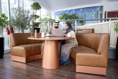 He uses a laptop to complete projects and tasks in the café, engaging in dialogue and discussion with colleagues, and spending enjoyable time with friends. An Emirati Gulf Arab man wearing a kandura and ghutrah sits with his female colleague in one of the cafés, displaying expressions of happiness and joy.