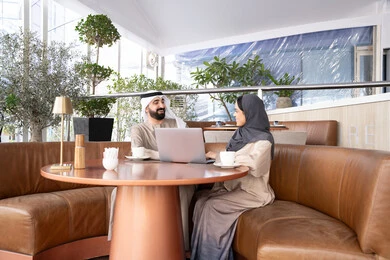 The dialogue and discussion between colleagues at work, using a laptop to complete projects and tasks in the café, spending enjoyable time with friends, an Emirati Gulf Arab man wearing a kandura and ghutrah sitting with his female colleague in one of the cafés, expressions of happiness and joy.