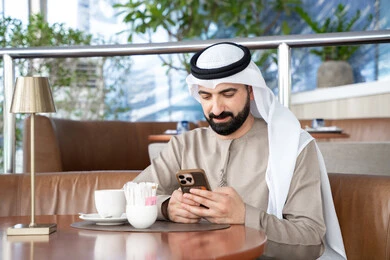 E-commerce and browsing mobile programs and applications, gestures of integration and focus, an Arab Gulf Emirati man wearing a kandura and ghutrah sitting in a café holding a mobile phone, the development of technology and technical devices, chatting with family and friends. E-commerce and browsing mobile programs and applications, gestures of integration and focus, an Arab Gulf Emirati man wearing a kandura and ghutrah sitting in a café holding a mobile phone, the development of technology and technical devices, chatting with family and friends.