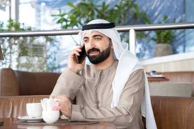 إجراء اتصال عبر الجوال، الرد على الاتصالات والمكالمات، رجل عربي خليجي اماراتي يرتدي الكندورة والغترة يجلس فى احد المقاهي يقوم باستخدام الهاتف المحمول ، مفهوم صلة الرحم و التواصل مع الأصدقاء والأقارب