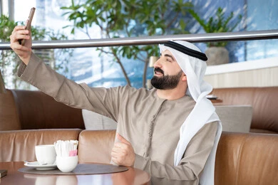 Video conversation through social media applications, taking commemorative selfies via mobile, using modern devices and technologies, an Arab Gulf Emirati man wearing the kandura and ghutrah is using a mobile phone, communicating with family and friends. Video conversation through social media applications, taking commemorative selfies via mobile, using modern devices and technologies, an Arab Gulf Emirati man wearing the kandura and ghutrah is using a mobile phone, communicating with family and friends.