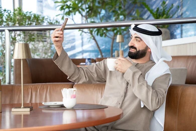 اجراء مكالمة فيديو عبر تطبيقات ومواقع السوشال ميديا، التقاط صور سيلفي تذكارية عبر الجوال, استخدام الأجهزة والتقنيات الحديثة، رجل عربي خليجي اماراتي يرتدي الكندورة والغترة يقوم باستخدام الهاتف المحمول، التواصل مع الأهل والأصدقاء، الاستمتاع بتناول المشروب المفضل