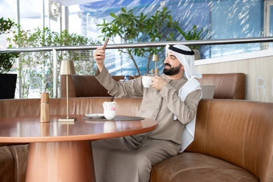 التقاط صور سيلفي تذكارية عبر الجوال, استخدام الأجهزة والتقنيات الحديثة، اجراء مكالمة فيديو عبر تطبيقات ومواقع السوشال ميديا، رجل عربي خليجي اماراتي يرتدي الكندورة والغترة يقوم باستخدام الهاتف المحمول، التواصل مع الأهل والأصدقاء، الاستمتاع بتناول المشروب المفضل