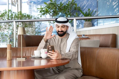 Exchanging news with family and friends, making a video call using a mobile phone, raising the palm of the hand is a sign of greeting and responding to peace, a portrait of an Emirati Gulf Arab man wearing a kandura and ghutrah sitting in a café communicating with relatives via mobile, expressions of joy and happiness.