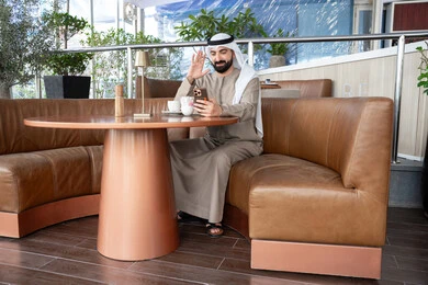 رفع كف اليد دليل على القاء التحية ورد السلام, إجراء مكالمة فيديو باستخدام الموبايل، تبادل الأخبار مع الأهل والأصدقاء، بورتريه لرجل عربي خليجي اماراتي يرتدي الكندورة و الغتره يجلس فى احد المقاهي يقوم بالتواصل مع الأقارب عبر الجوال، تعابير الفرح والسرور