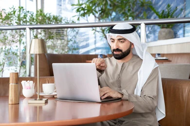 Pointing to the computer screen, typing on the keyboard, using modern and advanced technical devices to complete projects in the café, working remotely, a smiling Emirati Gulf Arab man wearing a kandura and a white ghutrah sitting in the café and using a laptop with gestures of engagement and focus.