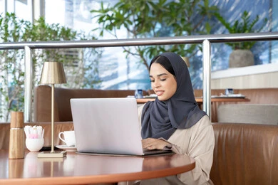 Browsing the internet and social media, video calls online, using modern devices and technologies, an Arab Gulf Emirati woman wearing an abaya and hijab using a laptop, a Saudi woman completing work tasks remotely, gestures of happiness and joy.