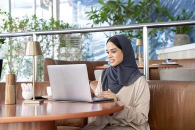 Conducting video calls, using modern devices and technologies, an Emirati Gulf Arab woman wearing an abaya and hijab using a laptop, a Saudi woman completing work tasks remotely, browsing the internet and social media online, gestures of happiness and joy.