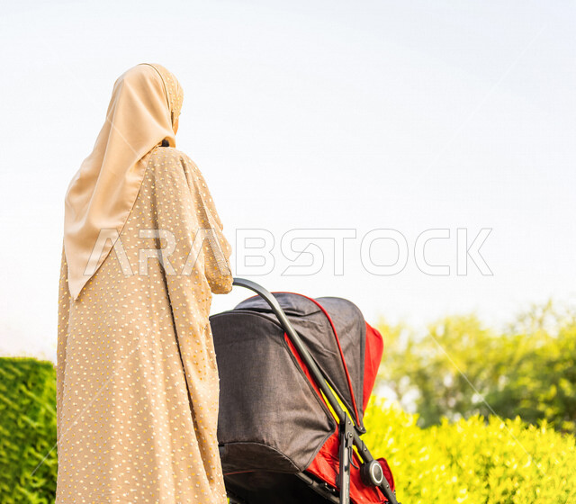 زاوية من الخلف لأم خليجية سعودية تمشي مع طفلها في عربة الأطفال في إحدى حدائق المملكة العربية السعودية ، مفهوم الأسرة السعيدة ، قضاء أوقات عائلية ممتعة ، أجواء عائلية ، عطلة نهاية الأسبوع ، إم وطفلها في رحلة ترفيهية ، جمال المناظر الطبيعية في السعودية