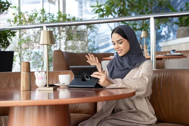 A Saudi woman is completing work tasks remotely using a tablet, an Emirati Gulf Arab woman wearing a hijab and abaya is sitting in an outdoor café, chatting and conversing through video calls, gestures of happiness and joy, sipping her favorite drink.