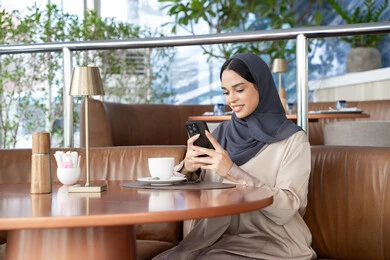 احتساء المشروب المفضل، تبادل الأحاديث المسليه عبر الجوال، الدردشة و التواصل الاجتماعي، امراة عربية خليجية اماراتية محجبة ترتدي العباءة تجلس على الأريكة في إحدى المقاهي الراقية تستمع بلحظات الهدوء والراحة ، سيده سعودية تقوم بتصفح المواقع عبر الانترنت