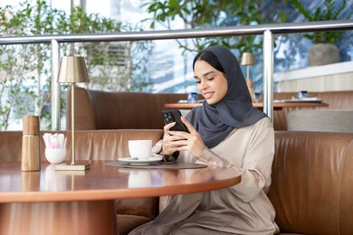 تبادل الأحاديث المسليه عبر الجوال، الدردشة و التواصل الاجتماعي، احتساء المشروب المفضل، امراة عربية خليجية اماراتية محجبة ترتدي العباءة تجلس على الأريكة في إحدى المقاهي الراقية تستمع بلحظات الهدوء والراحة ، سيده سعودية تقوم بتصفح المواقع عبر الانترنت