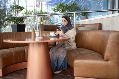 الدردشة و التواصل الاجتماعي، تبادل الأحاديث المسليه عبر الجوال، احتساء المشروب المفضل، امراة عربية خليجية اماراتية محجبة ترتدي العباءة تجلس على الأريكة في إحدى المقاهي الراقية تستمع بلحظات الهدوء والراحة ، سيده سعودية تقوم بتصفح المواقع عبر الانترنت