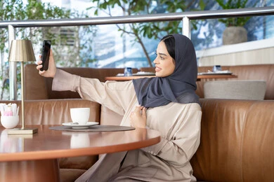 A Gulf Arab Emirati woman in a hijab wearing an abaya sits on a sofa in a high-end café, enjoying moments of calm and relaxation, taking selfies, connecting with family and friends, using modern devices and technologies, sipping her favorite drink, and a Saudi woman making a video call via her mobile phone.