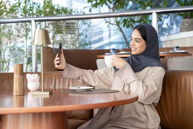 التواصل مع الأهل والأصدقاء، استخدام الأجهزة والتقنيات الحديثة، احتساء المشروب المفضل، امراة عربية خليجية اماراتية محجبة ترتدي العباءة تجلس على الأريكة في إحدى المقاهي الراقية تستمع بلحظات الهدوء والراحة ، سيده سعودية تقوم باجراء مكالمة فيديو عبر الهاتف المحمول، التقاط صور السيلفي