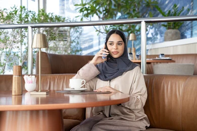 النقاش وتبادل الأخبار مع الأصدقاء والعائلة، اجراء مكالمة هاتفية عبر الجوال، استخدام جهاز تقني حديث ومتطور، امراة عربية خليجية اماراتية ترتدي العباءة والحجاب تجلس فى احد الكافيهات تمسك بيدها الهاتف المحمول، شابة سعودية تقوم بقضاء وقت ممتع في الخارج