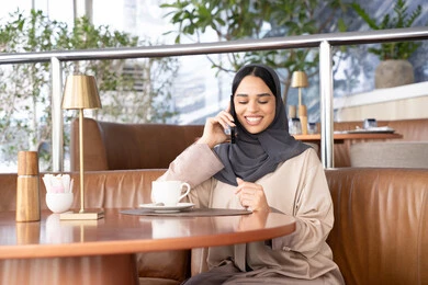 مشاركة الأخبار والحوارات مع العائلة والأصدقاء، اجراء مكالمة هاتفية عبر الجوال، استخدام جهاز تقني حديث ومتطور، امراة عربية خليجية اماراتية ترتدي العباءة والحجاب تجلس فى احد الكافيهات تمسك بيدها الهاتف المحمول، شابة سعودية تقوم بقضاء وقت ممتع في الخارج