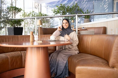 استخدام جهاز تقني حديث ومتطور، تبادل الأحاديث والأخبار مع الأهل والأصدقاء، اجراء مكالمة هاتفية عبر الجوال، امراة عربية خليجية اماراتية ترتدي العباءة والحجاب تجلس فى احد الكافيهات تمسك بيدها الهاتف المحمول، شابة سعودية تقوم بقضاء وقت ممتع في الخارج