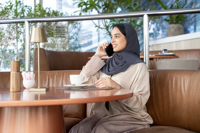 تبادل الأحاديث والأخبار مع الأهل والأصدقاء، اجراء مكالمة هاتفية عبر الجوال، امراة عربية خليجية اماراتية ترتدي العباءة والحجاب تجلس فى احد الكافيهات تمسك بيدها الهاتف المحمول، استخدام جهاز تقني حديث ومتطور، شابة سعودية تقوم بقضاء وقت ممتع في الخارج