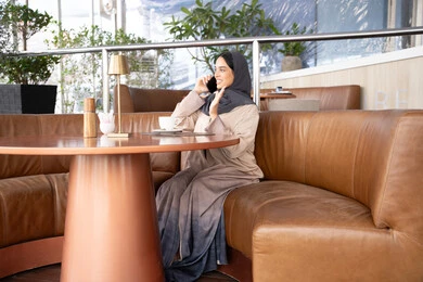 Making a phone call via mobile, exchanging conversations and news with family and friends, an Arab Gulf Emirati woman wearing an abaya and hijab sitting in one of the cafes holding a mobile phone in her hand, using a modern and advanced technological device, a Saudi young woman enjoying her time outside.