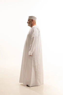 النظر إلى اشيء ما بإيماءات السرور والبشاشة، مفهوم الرجولة والوقار، الاهتمام بالمظهر الخارجي، الثقة بالنفس والوقوف بشكل مستقيم، بورتريه من الجانب لرجل كبير بالسن عربي خليجي عماني يرتدي الدشداشة التقليدية والكمه العمانية، الجسم بالطول الكامل، خلفية بيضاء