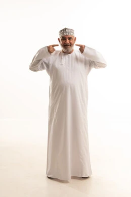 الوقوف باستقامة والنظر إلى الكاميرا, استخدام قشور فينير من الخزف لصنع ابتسامة بيضاء ناصعه، أهمية متابعة طبيب الاسنان، العناية والتجميل والتبيض، بورتريه لرجل مسن عربي خليجي عماني يرتدي الدشداشه والكمه يشير بيديه إلى أسنانه, صورة الجسم بالكامل, خلفية بيضاء