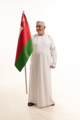 الفخر والاعتزاز بالانتماء لسلطنة عمان، تقديم الولاء للراية العمانية بمشاعر العز والكرامة، بورتريه لرجل شايب عربي خليجي عماني يرتدي الدشداشة والكمه التقليدية يحمل العلم العماني  بكل حب يقوم بالنظر للعلم، الجسم بالطول الكامل، خلفية بيضاء