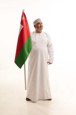 Presenting loyalty to the Omani flag with feelings of pride and dignity, pride and honor in belonging to the Sultanate of Oman, a portrait of an elderly Arab Gulf Omani man wearing the traditional dishdasha and kumma, holding the Omani flag with love while looking at something, full-body, white background.
