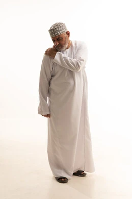 إيماءات الألم في الأكتاف، التهاب العضلات والمفاصل، الإحساس بالتعب والإرهاق, بورتريه لرجل مسن عربي عماني خليجي يرتدي الدشداشة والكمه يضع يده على كتفه بتعابير الوجع، الحاجه لوضع خطة علاجية, طلب المساعده للذهاب الى المستشفى، صورة الجسم بالكامل، خلفية بيضاء