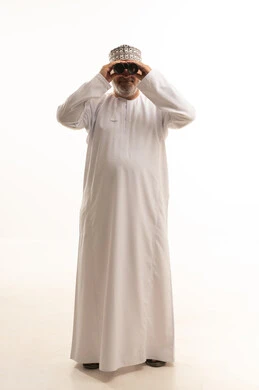 رؤية الاشياء البعيدة و تقريبها, ايماءات الاستمتاع و السرور, حب الاستكشاف و المغامرة, بورتريه لشاب عربي خليجي عماني يرتدي الدشداشة و الكمه مبتسم يحمل بيده منظار ثنائي العينين و يينظر الى شيئ ما, صورة الجسم بالطول الكامل, خلفية بيضاء