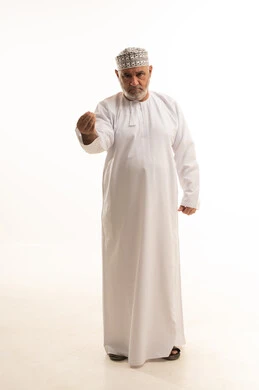 مفهوم الصبر والتريث، التوعد والتهديد لشخص ما، طلب التمهل في شيء ما، إشارة اليد تدل على التروي، بورتريه مقرب لرجل شايب عربي خليجي عماني يرتدي الدشداشة والكمه العمانية التقليدية يقف بشكل مستقيم وينظر إلى الكاميرا بإيماءات السرور والبشاشة، خلفية بيضاء
