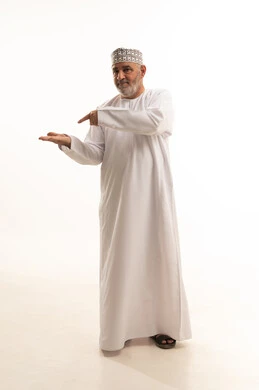 Pointing to something in the palm of the hand, standing and looking at the camera with gestures of cheerfulness, paying attention to the external appearance, a portrait of an elderly Arab Gulf Omani man wearing a dishdasha and kumma holding an item and pointing to it with his index finger, full body, white background.