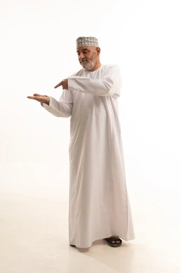In the palm of the hand, pointing to something, standing and looking at the camera with gestures of cheerfulness, paying attention to the external appearance, a portrait of an elderly Arab Gulf Omani man wearing a dishdasha and kumma, holding an item and pointing to it with his index finger, full-body, white background.
