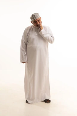 المعاناة من تسوس الأسنان، أوجاع التهابات اللثة وأمراض الفم، الحاجة لزيارة العيادة السنية، بورتريه لرجل شايب عربي خليجي عماني يرتدي الدشداشة والكمه العمانية يضع يده على خده ويغمض عينيه بإيماءات الألم الشديد، الجسم بالطول الكامل، خلفية بيضاء