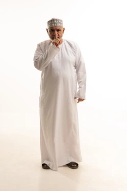 الأمر بعدم الازعاج والسكوت والصمت، بورتريه لشاب عربي خليجي عماني مبتسم يرتدي الدشداشة والكمه يضع اصبع السبابة على فمه ينظر الى الكاميرا بإيماءات التوقف عن الكلام وطلب خفض الصوت، تعابير الجدية والحزم والغضب، صورة الجسم بالطول الكامل، خلفية بيضاء