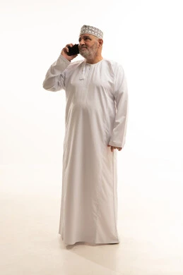 Gestures of happiness upon hearing good news, communication with family and friends, using modern and advanced technologies, a portrait of an elderly Arab Gulf Omani man wearing a dishdasha and an Omani kumma holding a mobile phone in his hand and making a phone call, full-body, white background.