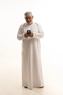 The use of modern and advanced technologies, communication with family and friends, gestures of happiness upon hearing good news, a portrait of an elderly Arab Gulf Omani man wearing a dishdasha and an Omani kumma holding a mobile phone in his hand and making a phone call, full-body, white background.