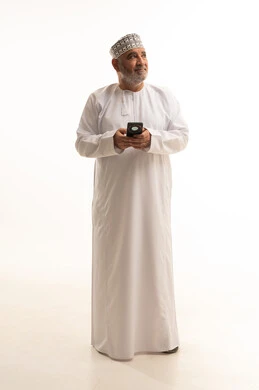 Looking at something above, using modern and advanced technologies, communicating with family and friends, a portrait of an elderly Arab Gulf Omani man wearing a dishdasha and an Omani kumma holding a mobile phone in his hand and having a phone conversation, full body, white background.
